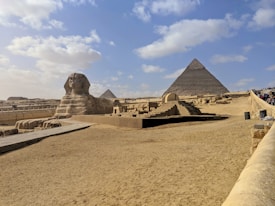 A vast desert landscape features the iconic Great Sphinx of Giza in the foreground and the Pyramid of Khafre in the background. The area is surrounded by sand and a clear blue sky, with scattered clouds adding depth to the scene. Tourists are visible along a path leading toward the structures, signifying its status as a historical and cultural landmark.