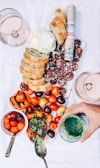 A charcuterie board is arranged with sliced bread, a roll of cured sausage, sliced salami, a variety of cherries, and grilled peaches topped with chimichurri sauce. A bowl of mixed melon balls and a spoon with green sauce accompany the spread. Two glasses of rosé wine are partially visible, and a hand is holding a jar of green sauce.