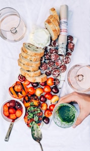 A charcuterie board is arranged with sliced bread, a roll of cured sausage, sliced salami, a variety of cherries, and grilled peaches topped with chimichurri sauce. A bowl of mixed melon balls and a spoon with green sauce accompany the spread. Two glasses of rosé wine are partially visible, and a hand is holding a jar of green sauce.