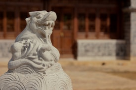 A detailed stone carving of a mythical creature, resembling a lion, is prominently displayed in the foreground. The intricate sculpture is mounted on a pedestal with wave-like patterns. In the background, there is a traditional wooden structure with ornate windows and a closed door, suggesting a historical or cultural setting.