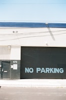 A large industrial building facade with a metal roll-up door displaying the words 'NO PARKING' in blue letters. The structure has a minimalistic design with a light beige wall and a dark grey or black door. There is a small metal door and a sign with the number 470 on a column next to the roll-up door. The area appears clean and well-maintained, with a clear blue sky above.