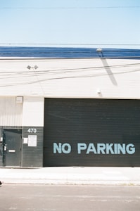 A large industrial building facade with a metal roll-up door displaying the words 'NO PARKING' in blue letters. The structure has a minimalistic design with a light beige wall and a dark grey or black door. There is a small metal door and a sign with the number 470 on a column next to the roll-up door. The area appears clean and well-maintained, with a clear blue sky above.