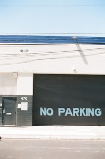 A large industrial building facade with a metal roll-up door displaying the words 'NO PARKING' in blue letters. The structure has a minimalistic design with a light beige wall and a dark grey or black door. There is a small metal door and a sign with the number 470 on a column next to the roll-up door. The area appears clean and well-maintained, with a clear blue sky above.