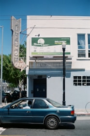 A blue car is parked on the street in front of a building with a large sign advertising for Mission Organic. The building is light blue with white trim and has a vintage-style vertical sign. A streetlamp is positioned near the car.