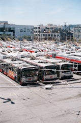 A large number of electric buses are parked in an expansive outdoor parking lot. Each bus has distinctive overhead trolley poles connected to power lines above. The background features modern buildings, some with distinct balconies and various architectural designs.