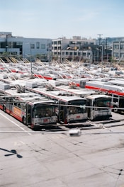 A large number of electric buses are parked in an expansive outdoor parking lot. Each bus has distinctive overhead trolley poles connected to power lines above. The background features modern buildings, some with distinct balconies and various architectural designs.