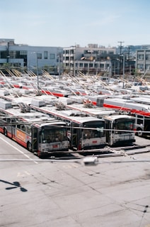 A large number of electric buses are parked in an expansive outdoor parking lot. Each bus has distinctive overhead trolley poles connected to power lines above. The background features modern buildings, some with distinct balconies and various architectural designs.