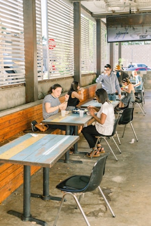 A group of people are sitting at outdoor tables in a casual setting with wooden benches and metal chairs. Some are engaged in conversation, while others appear to be looking at their phones. There is a large screen displaying a sports game mounted on the wall. The area has an open feel with horizontal slats allowing in sunlight.