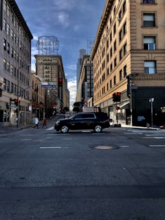 A city street intersection with tall buildings on either side. A black SUV is prominently in the middle of the intersection. People are visible walking on the sidewalks. The street signs indicate this is Los Angeles Street. Bright neon hotel signs are on the buildings, and a cloudy sky stretches above.