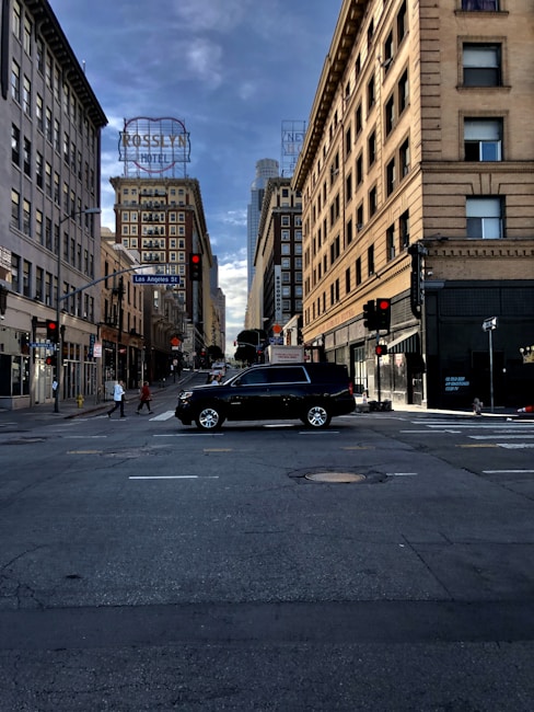 A city street intersection with tall buildings on either side. A black SUV is prominently in the middle of the intersection. People are visible walking on the sidewalks. The street signs indicate this is Los Angeles Street. Bright neon hotel signs are on the buildings, and a cloudy sky stretches above.