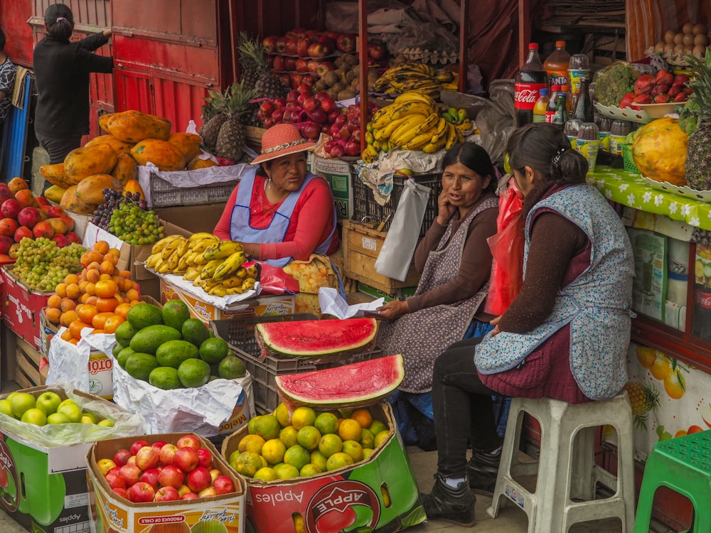 Drei Frauen sitzen hinter verschiedenen Früchten, die auf dem Bürgersteig ausgestellt sind