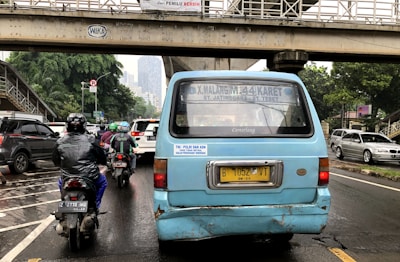 A busy urban street scene featuring a blue passenger van with distinct wear and tear on the back. Two motorcyclists are riding behind the van. The surrounding environment includes several cars, a pedestrian overpass, and urban greenery. The image depicts a typical traffic situation in a city.