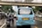 A busy urban street scene featuring a blue passenger van with distinct wear and tear on the back. Two motorcyclists are riding behind the van. The surrounding environment includes several cars, a pedestrian overpass, and urban greenery. The image depicts a typical traffic situation in a city.