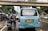 A busy urban street scene featuring a blue passenger van with distinct wear and tear on the back. Two motorcyclists are riding behind the van. The surrounding environment includes several cars, a pedestrian overpass, and urban greenery. The image depicts a typical traffic situation in a city.