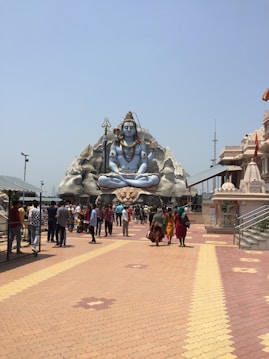 A large statue of a deity in a seated lotus position dominates the background, situated on a rocky structure. The deity is adorned with traditional ornaments, with a trident nearby. In the foreground, a group of people walks along a patterned pathway leading towards the statue. The surrounding architecture includes temple-like buildings, with bright skies overhead.