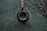 A dark, textured background is scattered with coffee beans, placing focus on a metallic container with a wooden handle, possibly a mate cup. A metal straw is inserted into the container, and nearby, thin branches with small, light-colored buds are arranged diagonally.