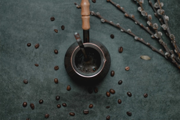 A dark, textured background is scattered with coffee beans, placing focus on a metallic container with a wooden handle, possibly a mate cup. A metal straw is inserted into the container, and nearby, thin branches with small, light-colored buds are arranged diagonally.