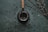 A dark, textured background is scattered with coffee beans, placing focus on a metallic container with a wooden handle, possibly a mate cup. A metal straw is inserted into the container, and nearby, thin branches with small, light-colored buds are arranged diagonally.