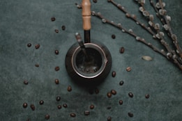 A dark, textured background is scattered with coffee beans, placing focus on a metallic container with a wooden handle, possibly a mate cup. A metal straw is inserted into the container, and nearby, thin branches with small, light-colored buds are arranged diagonally.