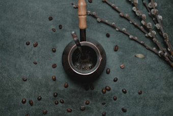 A dark, textured background is scattered with coffee beans, placing focus on a metallic container with a wooden handle, possibly a mate cup. A metal straw is inserted into the container, and nearby, thin branches with small, light-colored buds are arranged diagonally.