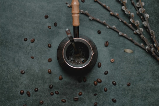 A dark, textured background is scattered with coffee beans, placing focus on a metallic container with a wooden handle, possibly a mate cup. A metal straw is inserted into the container, and nearby, thin branches with small, light-colored buds are arranged diagonally.
