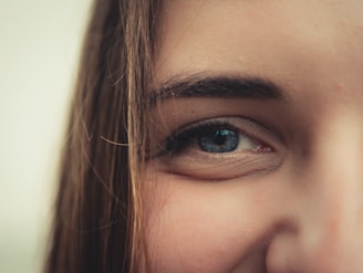 A close-up of a person's face focusing on the right eye and surrounding area, highlighting the details of eyelashes, eyebrow, and skin texture. The image emphasizes the blue color of the eye with strands of brown hair partially covering the forehead.