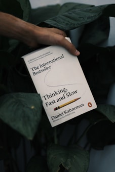A hand holds a book titled 'Thinking, Fast and Slow' against a backdrop of dark green leaves. The book cover is predominantly white with black and yellow text, featuring a pencil graphic.