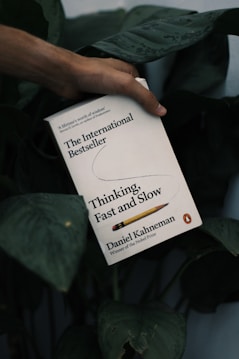 A hand holds a book titled 'Thinking, Fast and Slow' against a backdrop of dark green leaves. The book cover is predominantly white with black and yellow text, featuring a pencil graphic.