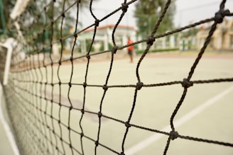 A close-up view of a sports net with intricate knots, set against the blurred background of an outdoor tennis or volleyball court. The court surface is visible with painted lines, and some indistinct structures can be seen in the distance.