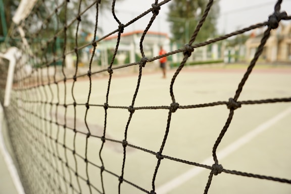 A close-up view of a sports net with intricate knots, set against the blurred background of an outdoor tennis or volleyball court. The court surface is visible with painted lines, and some indistinct structures can be seen in the distance.