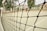 A close-up view of a sports net with intricate knots, set against the blurred background of an outdoor tennis or volleyball court. The court surface is visible with painted lines, and some indistinct structures can be seen in the distance.