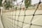 A close-up view of a sports net with intricate knots, set against the blurred background of an outdoor tennis or volleyball court. The court surface is visible with painted lines, and some indistinct structures can be seen in the distance.