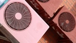 Several reddish-pink air conditioning units are mounted on a brick wall. The units show signs of wear with some visible rust and discoloration. Each unit features a circular fan and protective grille on the front.
