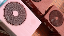 Several reddish-pink air conditioning units are mounted on a brick wall. The units show signs of wear with some visible rust and discoloration. Each unit features a circular fan and protective grille on the front.
