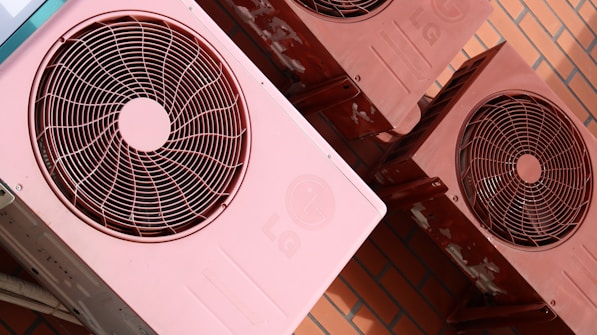 Several reddish-pink air conditioning units are mounted on a brick wall. The units show signs of wear with some visible rust and discoloration. Each unit features a circular fan and protective grille on the front.