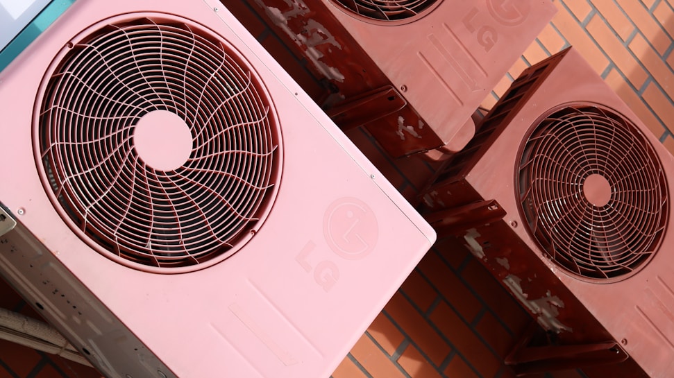 Several reddish-pink air conditioning units are mounted on a brick wall. The units show signs of wear with some visible rust and discoloration. Each unit features a circular fan and protective grille on the front.