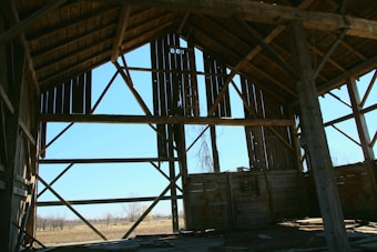 An old, wooden barn with its framing exposed. The structure features large vertical and diagonal beams, creating a geometric pattern against the bright blue sky. Sunlight filters through gaps in the wood, casting shadows on the interior dirt floor.