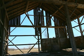 An old, wooden barn with its framing exposed. The structure features large vertical and diagonal beams, creating a geometric pattern against the bright blue sky. Sunlight filters through gaps in the wood, casting shadows on the interior dirt floor.