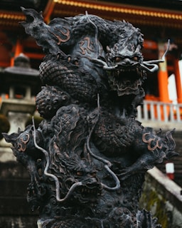 A detailed and intricately carved dark stone statue of a mythical creature, resembling a dragon. The statue is positioned in front of a traditional structure with vibrant orange and white elements, which could be a temple or shrine. The creature has an imposing presence, with its aggressive posture, open mouth, and meticulously patterned scales.
