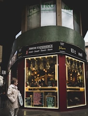 A street scene featuring a corner storefront with a sign advertising 'AMON EXPERIENCE.' The store has illuminated displays with hanging cured meats. Menus and chalkboards are visible outside the store. A person wearing a beige coat holds a shopping bag, and a child walks nearby on the sidewalk.