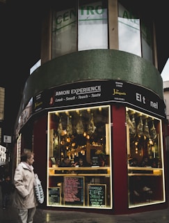 A street scene featuring a corner storefront with a sign advertising 'AMON EXPERIENCE.' The store has illuminated displays with hanging cured meats. Menus and chalkboards are visible outside the store. A person wearing a beige coat holds a shopping bag, and a child walks nearby on the sidewalk.
