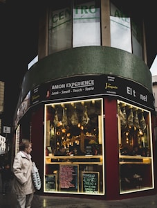 A street scene featuring a corner storefront with a sign advertising 'AMON EXPERIENCE.' The store has illuminated displays with hanging cured meats. Menus and chalkboards are visible outside the store. A person wearing a beige coat holds a shopping bag, and a child walks nearby on the sidewalk.