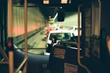 The interior of a bus looking out into a traffic-filled tunnel. Vehicles are lined up with brake lights glowing, and the dimly lit tunnel features reflective walls. The bus interior includes visible bars, a windshield, and signage with welcoming or instructional messages.