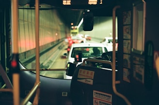 The interior of a bus looking out into a traffic-filled tunnel. Vehicles are lined up with brake lights glowing, and the dimly lit tunnel features reflective walls. The bus interior includes visible bars, a windshield, and signage with welcoming or instructional messages.
