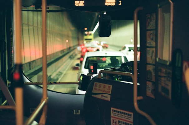 The interior of a bus looking out into a traffic-filled tunnel. Vehicles are lined up with brake lights glowing, and the dimly lit tunnel features reflective walls. The bus interior includes visible bars, a windshield, and signage with welcoming or instructional messages.