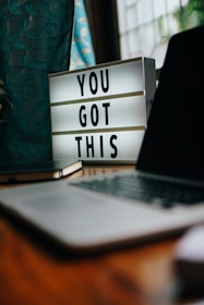 A motivational lightbox displaying the message 'YOU GOT THIS' sits on a wooden desk. Nearby, a closed laptop and a notebook are visible. The setting is by a window with teal curtains that have a leaf pattern.