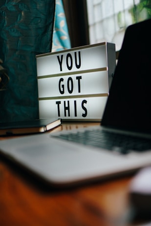 A motivational lightbox displaying the message 'YOU GOT THIS' sits on a wooden desk. Nearby, a closed laptop and a notebook are visible. The setting is by a window with teal curtains that have a leaf pattern.