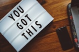 A lightbox with the motivational phrase 'YOU GOT THIS' is on a wooden surface. Nearby is a black smartphone and a laptop with a floral design on its sleeve.