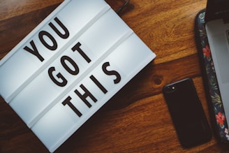 A lightbox with the motivational phrase 'YOU GOT THIS' is on a wooden surface. Nearby is a black smartphone and a laptop with a floral design on its sleeve.