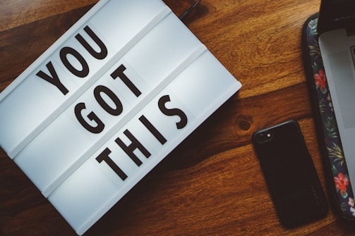 A lightbox with the motivational phrase 'YOU GOT THIS' is on a wooden surface. Nearby is a black smartphone and a laptop with a floral design on its sleeve.